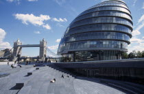 City Hall exterior view of the curved architecture with Tower Bridge in the distance and people sitting on the steps in the foreground