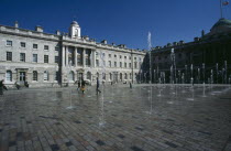 Somerset House and courtyard with rows of fountains spouting high from the ground with people walking between them. Flag flying at half mast in the distance.