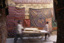Jaisalmer Fort. Two men pushing a cart through a narrow street with textile rugs hanging from the walls