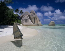 Large rock formations on sandy beach with clear shallow water and palmtrees growing along coastline