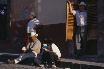 Street scene with man and young boys in conversation sitting on pavement and man and woman standing behind.