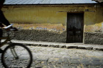 A backstreet in the outskitrs of the Guatemalan town of Xela. A characteristic but crumbling house sits baking in the sun  infront lies a dishevelled pavement and a dry dusty cobbled road. The house i...
