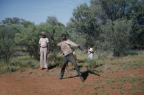 Aborigine man throwing a Boomerang with a man and children standing behind him looking on