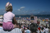 View over crowds on the beach viewing Punch and Judy show with girl in pink sitting on man shoulders in the foreground