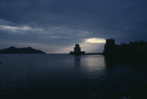 View across the sea towards fortress ruins lit by evening sun rays coming through a break in the clouds.