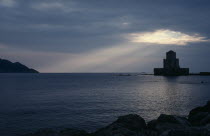 View across the sea towards fortress ruins lit by evening sun rays coming through a break in the clouds.