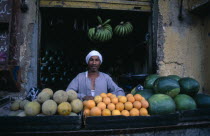 Fruit vendor outside shop selling green bananas  water melons and oranges