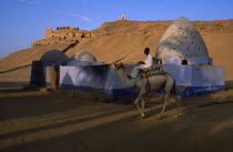 Egypt, Upper Egypt, Aswan, Man on camel rides past Beehive Mausoleum with Tomb of the Nobles on hilltop behind.