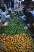 Fruit and vegetable market with vendors and customers inspecting the produce of green chilli peppers and oranges