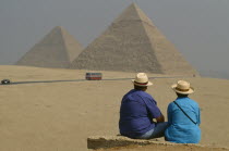 The Pyramids with two tourists seated in foreground. Bus on road.