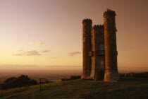 Broadway Tower seen in golden light with people standing on grass in the grounds behind towerDesigned to resemble a mock castle on a beacon hill. Standing 1024 feet 312m above sea level
