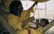 Mother and baby son in a  fique  / cactus fibre hammock in their desert home. Mother with black fungal facial paint to protect against strong wind and sun. She sews a  faja  / woven woollen belt for h...