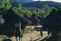 Parque Nacional. Kogi Wiwa leader riding his horse through Avingue village between mud-wattle walled and grass thatched homes with children playing   banana palms and cultivation plots surrounding vil...