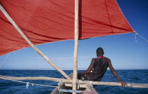 Ifaty Beach. Young fisherman sitting on bow of red sail Pirogue boatA pirogue is a small flat hulled boat of a design associated particularly with West African fishermen