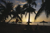 Ifaty Beach at sunset with view through palm trees and straw sun shades towards the sea with people on the sand in silhouette
