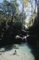 Trees surrounding a waterfall running over rocks into a natural fresh pool with a woman swimming in the clear water