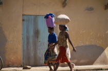 Two women wearing colourful clothing walking along a street next to a building one carrying a shopping basket on her head and the other woman carrying a bag of grain