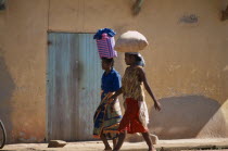 Two women wearing colourful clothing walking along a street next to a building one carrying a shopping basket on her head and the other woman carrying a bag of grain