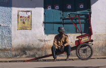 Man sitting on his rickshaw awaiting a fare at the side of the road with a building behind