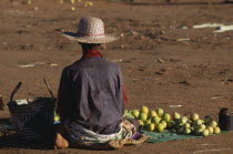 Woman sat on ground with her back facing the lens wearing straw hat and Chicago Bulls basket ball shirt selling fruit