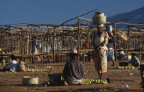 Two women selling fruit at a sunday market place with empty stalls one woman sat on ground with her back turned and one woman standing carrying wicker baskets on her head facing lens
