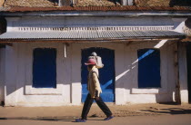 Boy wearing a red cap carrying a bag on top of his shoulder walking past a white washed building with a blue door and shutters