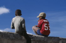 Two boys sitting on a wall one wearing a red baseball shirt and cap