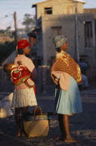 Road to Fianarantsoa. Two women one carrying a baby in a sling on her back wearing colourful clothing standing waiting for a lift with golden sunlight shining onto them
