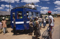 Road to Antsirabe. Minibus at a roadside stopping point with a woman and children selling pineapple from bowls to the passengers onboard and a man wearing a helmet  ona bicycle in the foreground.
