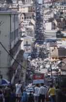 Downtown area. People walking down towards a busy road running between buildings and shopfronts filled with people and cars