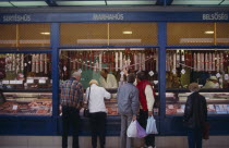 Tourists buying salami from butchers with counter open on to street.