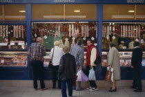 Tourists buying salami from butchers with counter open on to street.