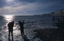 People in silhouette paddling at the waters edge on pebbled beachBeaches European Great Britain Northern Europe Resort Sand Sandy Seaside Shore Tourism UK United Kingdom British Isles