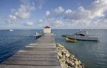 Wooden jetty stretching out to sea with a man sat next to a hut at the end of the jetty with boats moored on the water nearby