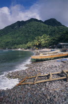 Scotts Head fishing village. View across stoney beach lined with wooden fishing boats and huts towards palmtrees and tree covered hillside