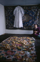 A white wedding dress hangs on the wall in front of a rug  and a large display of food is laid out awaiting guests from a village wedding
