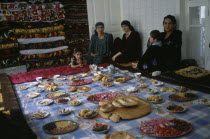 Three women and two children sit by a large display of food laid out  awaiting the guests from the village wedding.
