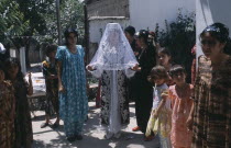 The bride in a white dress  veil and decorated jacket surrounded by guests at a village wedding.