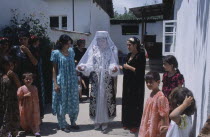 The bride wearing a veil and highly decorated jacket  surrounded by guests at a village wedding.