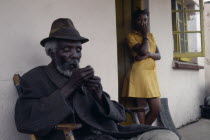 Portrait of elderly seated man lighting pipe with girl standing in doorway behind.