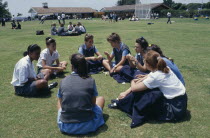 Mixed race group of schoolgirls sitting on playing fields during break.