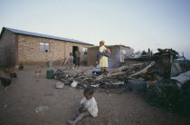 Mother and children outside their home with chickens and cooking fire.  Young child playing in the dirt in the foreground.family