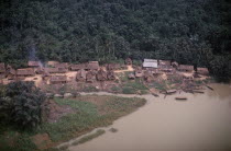 Aerial view over thatched village on river bank beside area of tropical forest.