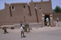 Traditional mud Hausa dwelling with child standing with up-stretched arms outside and man entering through open doorway.