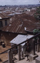 View across corrugated tin rooftops of city houses with child carrying water jar in the foreground.