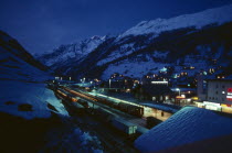 Ski resort chalets and train station in snow covered mountain landscape at night with lights from windows and platform.