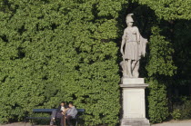 Schonbrunn Gardens.  Classical statue against hedge with two visitors sitting on bench at one side.
