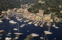 Aerial view over harbour with moored yachts and pastel coloured town houses with tree covered hillside behind.Colored