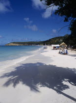 View along quiet sandy beach towards sunbathers and beach stall with headland beyond.  Shadow from palm tree in the foreground.