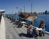 Corniche. Restaurant Dhow by seam moored boats with two woman sat on table and chairs in the foreground United Arab Emirates
